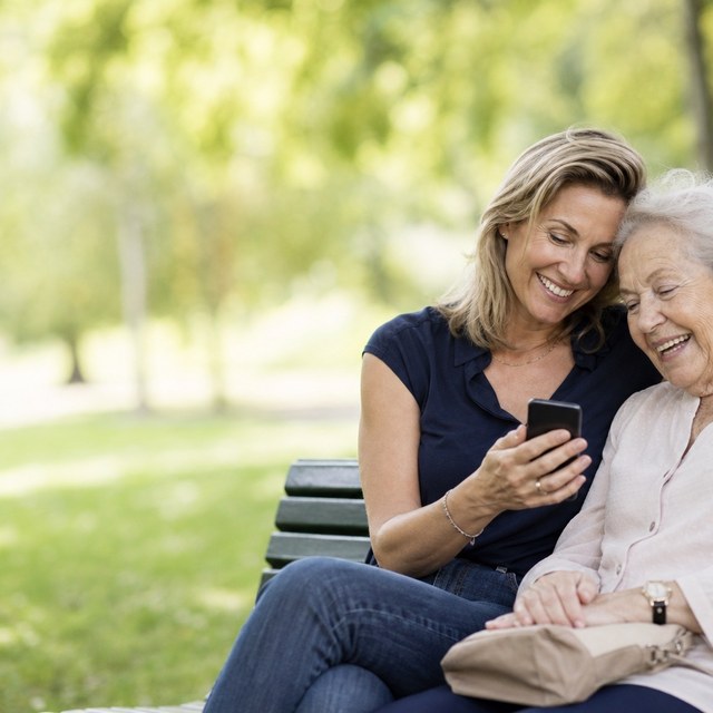 A woman and her elderly mother on a park bench, heads together over one phone, both smiling.